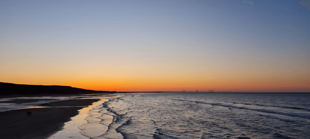 View looking along the beach and sea, with the tide coming in, beneath an orange sunset and blue sky